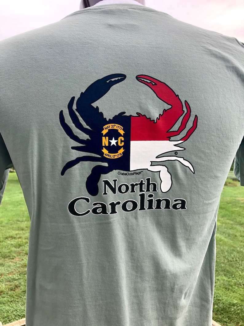 May include: Sage green t-shirt featuring a crab design with the North Carolina state flag colors. The crab's claws are navy and red, with the state seal in the center. The words "North Carolina" are printed below the crab.