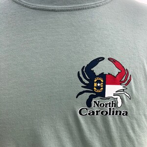 May include: Sage green t-shirt featuring a crab design with the North Carolina state flag incorporated into the crab's claws. The words "North Carolina" are printed below the crab in a stylized font.