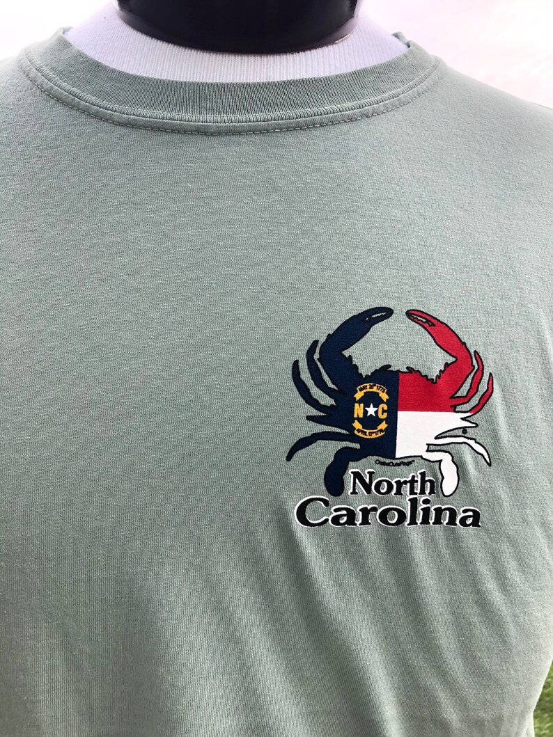 May include: Sage green t-shirt featuring a crab design with the North Carolina state flag incorporated into the crab's claws. The words "North Carolina" are printed below the crab in a stylized font.
