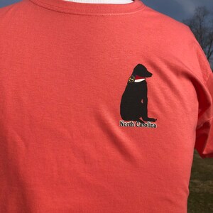 K&ouml;nnte beinhalten: Korallenfarbenes T-Shirt mit einer schwarzen Hunde-Grafik, wei&szlig;em Kragen und Abzeichen. Der Text "North Carolina" befindet sich unter dem Hund. Das T-Shirt wird auf einer Schaufensterpuppe pr&auml;sentiert.