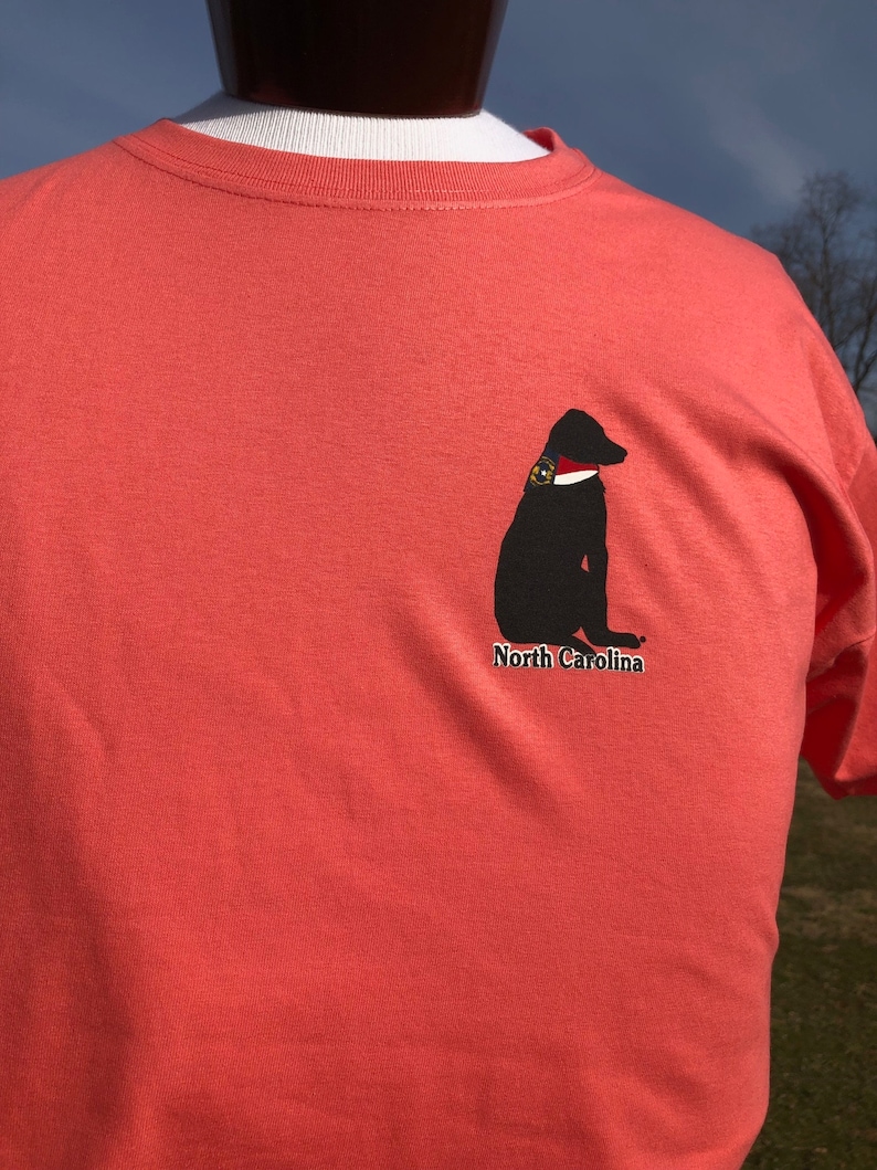 K&ouml;nnte beinhalten: Korallenfarbenes T-Shirt mit einer schwarzen Hunde-Grafik, wei&szlig;em Kragen und Abzeichen. Der Text "North Carolina" befindet sich unter dem Hund. Das T-Shirt wird auf einer Schaufensterpuppe pr&auml;sentiert.