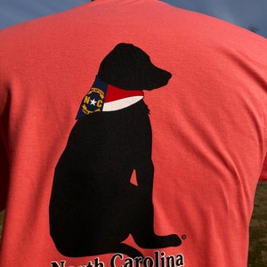 K&ouml;nnte beinhalten: Korallenfarbenes T-Shirt mit einer schwarzen Silhouette eines Hundes, der ein Halstuch im Stil der Flagge von North Carolina tr&auml;gt. Die Worte "North Carolina" sind unten in Wei&szlig; aufgedruckt. Das Halstuch des Hundes hat einen blauen Hintergrund mit einem wei&szlig;en Stern und den Buchstaben "N C".