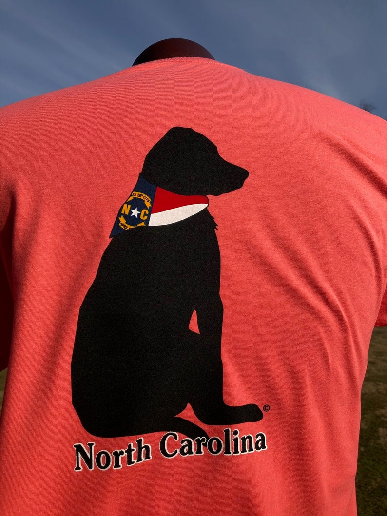 K&ouml;nnte beinhalten: Korallenfarbenes T-Shirt mit einer schwarzen Silhouette eines Hundes, der ein Halstuch im Stil der Flagge von North Carolina tr&auml;gt. Die Worte "North Carolina" sind unten in Wei&szlig; aufgedruckt. Das Halstuch des Hundes hat einen blauen Hintergrund mit einem wei&szlig;en Stern und den Buchstaben "N C".