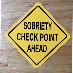 May include: A yellow diamond sign with the words "SOBRIETY CHECK POINT AHEAD" in black. The sign has a black border and is placed on a wooden surface.