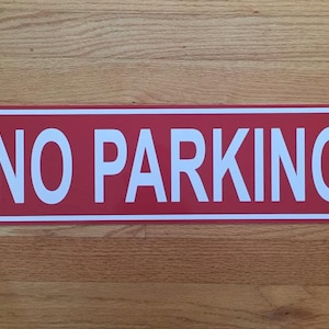 May include: A rectangular red metal sign with white text that reads "NO PARKING". The sign has a white border and is displayed on a wooden surface.