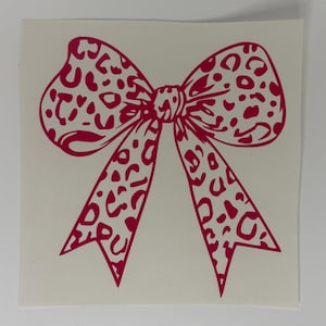 May include: A red bow sticker with a leopard print pattern. The bow is centered on a white background. The bow's design features a series of irregular spots, creating a bold and eye-catching aesthetic. Ideal for crafting and decorating.