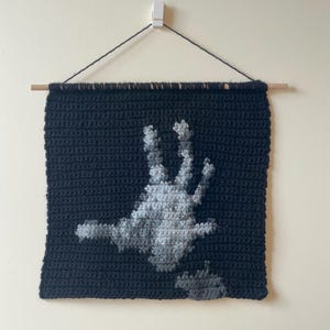 May include: A handmade, dark blue crochet wall hanging featuring a grayscale handprint design. The artwork is suspended from a wooden dowel with black string. The handprint is detailed with varying shades of gray.