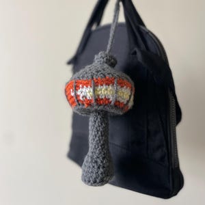 May include: A handmade crochet charm in the shape of a mushroom, featuring a gray stem and cap. The cap is decorated with orange, white, and yellow stripes. The charm is attached to a black bag by a gray string.