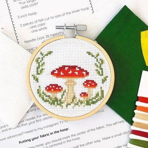 May include: A cross stitch embroidery kit featuring a mushroom design. The kit includes aida cloth, embroidery floss, a hoop, and felt. The instructions include a color pattern and symbols pattern for stitching.
