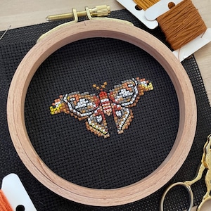 May include: A cross-stitch butterfly design in a wooden embroidery hoop. The butterfly is stitched with orange, yellow, white, and brown thread on black fabric. Also pictured are spools of orange thread and gold scissors.