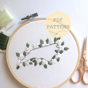 May include: A white embroidery hoop with a black outline of the state of North Carolina surrounded by white flowers and green leaves. The hoop is on a white surface with a pair of gold scissors and a spool of green thread.