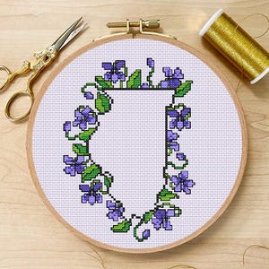 Può includere: Un telaio da ricamo a punto croce con la lettera "J" circondata da fiori viola e foglie verdi. Il ricamo è in un telaio di legno, con forbici e filo dorati. Lo sfondo è una superficie di legno chiaro.
