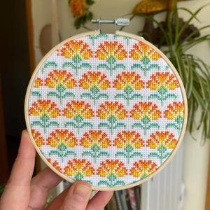 May include: A round cross-stitch embroidery hoop featuring a repeating floral pattern. The flowers are orange and yellow with green stems and leaves, set against a white background. The wooden hoop is held by a hand.