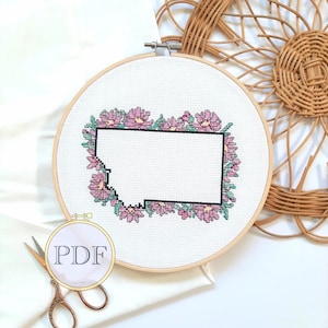 Puede incluir: Un aro de bordado de punto de cruz blanco con un borde floral rosa alrededor del contorno del estado de Montana. El aro está sobre una superficie de tela blanca.