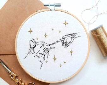 May include: Embroidered hoop art featuring a cat looking up at a skeletal hand reaching out with a finger. The background is white with gold glitter stars.