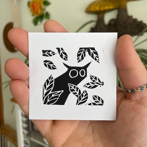May include: A black and white linocut print featuring a stylized owl with large eyes, surrounded by leaves. The owl is the focal point, set against a white background. The print has a handmade, artistic feel.