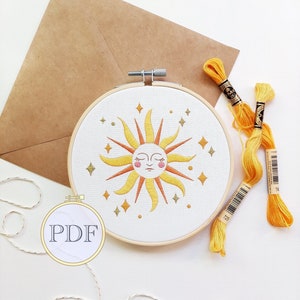 May include: A white embroidery hoop with a yellow and orange sun with a face and closed eyes. The sun is surrounded by gold stars. The hoop is on a white surface with two spools of yellow embroidery floss.