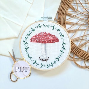 May include: A cross-stitch embroidery hoop with a red and white mushroom design surrounded by green leaves. The hoop is made of wood and has a white background.