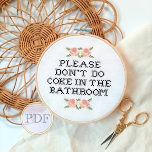 以下が含まれることがあります： 白い刺繍枠に「PLEASE DON'T DO COKE IN THE BATHROOM」という黒い文字が書かれています。枠は籐のバスケットと白い布で囲まれています。枠にはピンクの花が刺繍されています。