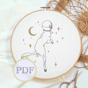 以下が含まれることがあります： 白い刺繍枠に、宇宙服を着た犬の黒い線画が描かれています。犬は金色のキラキラ光る星と三日月で囲まれています。枠には「PDF」という黒い文字が書かれた白いラベルが付いています。