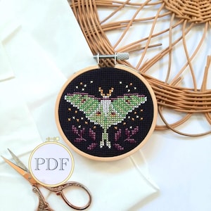 May include: A cross-stitch embroidery hoop with a black background and a green and white luna moth with purple floral accents.