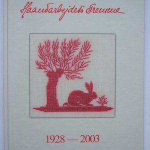 May include: A book with a cream-colored cover featuring red text and a cross-stitch design. The design includes a tree, a rabbit, and the years 1928-2003. The title is written in cursive at the top.