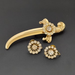 May include: A gold-toned vintage jewelry set featuring a brooch in the shape of a sword and two matching earrings. The brooch and earrings are adorned with clear, sparkling rhinestones arranged in a floral pattern. The set is displayed on a dark surface.
