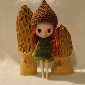 May include: A doll with red hair, blue eyes, and a green dress, wearing a brown knitted hat, stands between two carved wooden mushroom sculptures. The doll is the focal point, with the mushrooms providing a rustic backdrop.