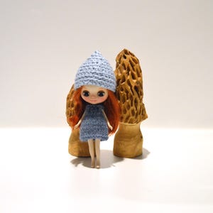 May include: A small doll with red hair and a blue knitted dress and hat. The doll is standing in front of two wooden mushroom sculptures. The background is plain white.