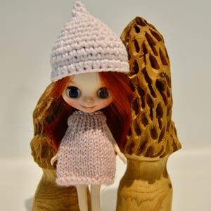 May include: A doll with red hair and blue eyes wearing a light pink knitted dress and matching hat. Two carved wooden mushroom sculptures flank the doll. The doll is standing on a white surface.