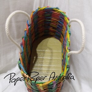 May include: A colorful woven paper tote bag with white rope handles. The bag is made of strips of paper in various colors, including red, orange, yellow, green, blue, purple, and pink. The bag is empty and the inside is visible. The text "Paper Paper Australia" is printed on the bottom of the bag.
