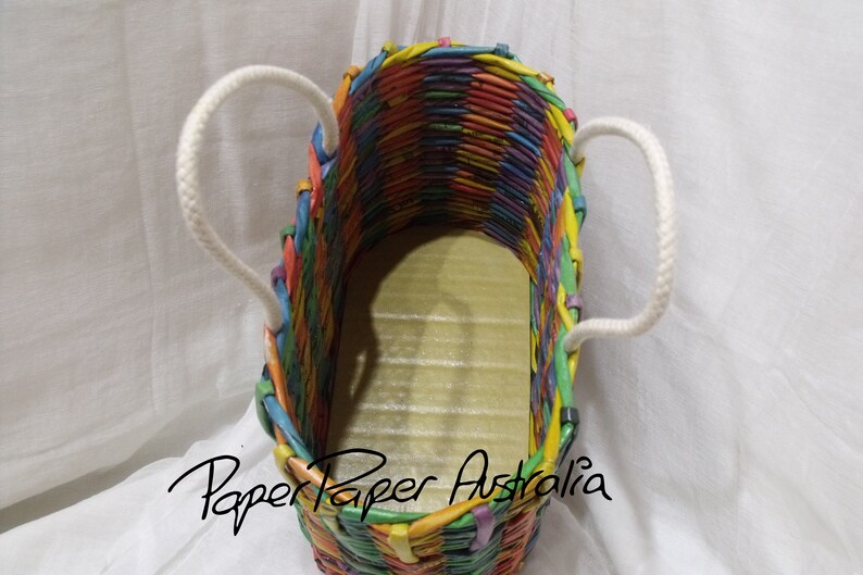 May include: A colorful woven paper tote bag with white rope handles. The bag is made of strips of paper in various colors, including red, orange, yellow, green, blue, purple, and pink. The bag is empty and the inside is visible. The text "Paper Paper Australia" is printed on the bottom of the bag.