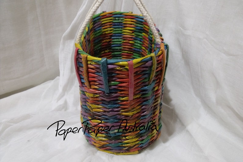 May include: A colorful woven basket with a white rope handle. The basket is made of paper and has a rainbow pattern.