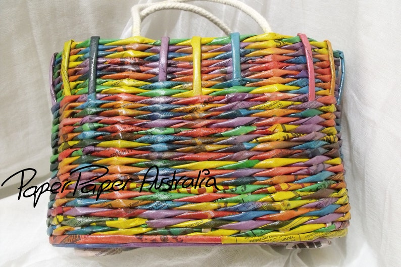 May include: A colorful woven tote bag made from recycled paper. The bag has a white handle and is woven with strips of paper in various colors, including yellow, orange, red, purple, blue, and green. The bag is labeled "Paper Paper Australia".