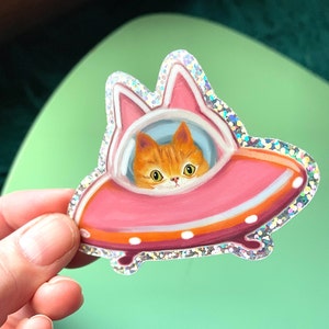 Cute and funny painterly orange kitty cat glitter sticker taking a spin in the abandoned pink alien flying saucer designed just for him-who wouldn’t?