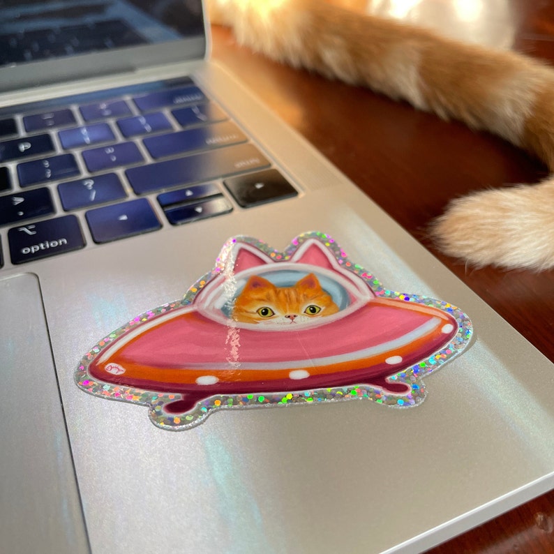 Cute and funny painterly orange kitty cat glitter sticker taking a spin in the abandoned pink alien flying saucer designed just for him-who wouldn’t?