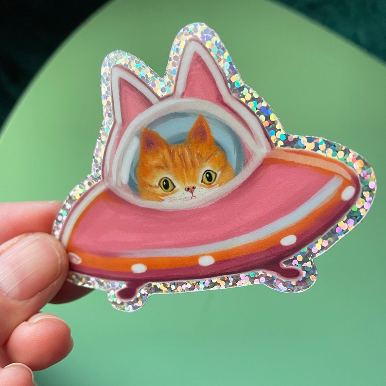 Cute and funny painterly orange kitty cat glitter sticker taking a spin in the abandoned pink alien flying saucer designed just for him-who wouldn’t?