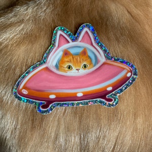 Cute and funny painterly orange kitty cat glitter sticker taking a spin in the abandoned pink alien flying saucer designed just for him-who wouldn’t?