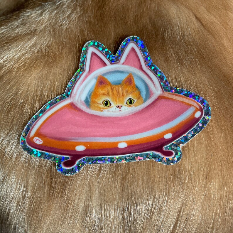 Cute and funny painterly orange kitty cat glitter sticker taking a spin in the abandoned pink alien flying saucer designed just for him-who wouldn’t?