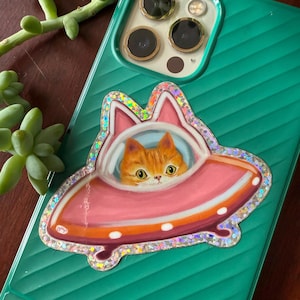 Cute and funny painterly orange kitty cat glitter sticker taking a spin in the abandoned pink alien flying saucer designed just for him-who wouldn’t?