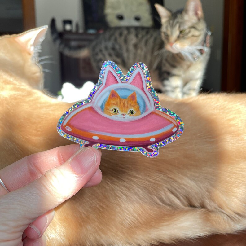 Cute and funny painterly orange kitty cat glitter sticker taking a spin in the abandoned pink alien flying saucer designed just for him-who wouldn’t?