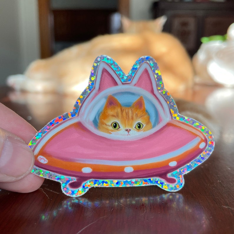 Cute and funny painterly orange kitty cat glitter sticker taking a spin in the abandoned pink alien flying saucer designed just for him-who wouldn’t?