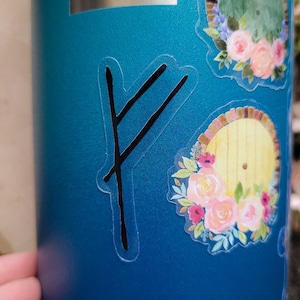 May include: A teal blue water bottle with a black vinyl sticker of a rune symbol. The rune symbol is a double line with a diagonal line across the top. The bottle also has two floral stickers with a yellow door in the center.