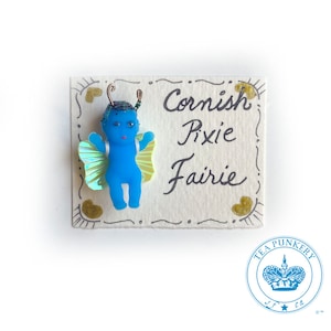 May include: A blue pixie figurine with iridescent wings is affixed to a cream-colored card. The card features the words "Cornish Pixie Fairie" in black script, with gold heart accents and a decorative border. A blue logo is in the bottom right corner.