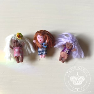 May include: Three small dolls with different hair colors and outfits are arranged in a row. The dolls have white, red, and purple hair. The dolls are wearing dresses in pink, blue, and gold colors. The dolls are miniature and appear to be handmade.