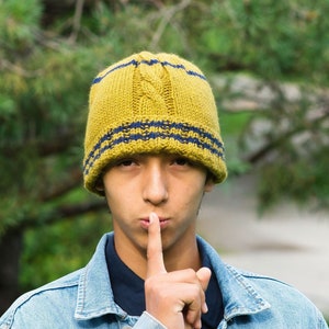 Peut inclure: Une jeune personne portant un bonnet en tricot jaune et bleu et une veste en jean bleue, avec son doigt sur ses lèvres, comme pour dire 'chut'.