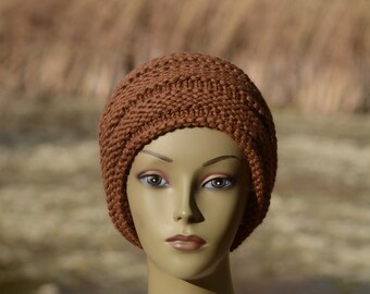 Puede incluir: Un gorro de punto marrón con un patrón de punto de cable. El gorro está en un maniquí que lleva una chal de punto blanco con cierre de botón.