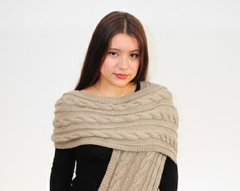 Può includere: Sciarpa beige a trecce drappeggiata sulle spalle, con un lungo pannello pendente. La sciarpa è realizzata in un materiale morbido e strutturato. La persona indossa un top nero a maniche lunghe e una gonna nera.