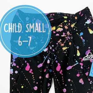 Könnte beinhalten: Schwarze Leggings mit einem mehrfarbigen Farbspritzer-Muster und einem passenden Haargummi. Die Leggings haben ein Etikett mit der Aufschrift CHILD SMALL 6-7. Das Design zeigt rosa, blaue, gelbe und violette Spritzer auf dem Stoff.