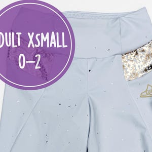 Könnte beinhalten: Hellblaue Leggings mit einem silber- und goldfarbenen Folienmuster. Die Leggings haben eine Tasche mit einem goldenen Paillettenmuster. Ein passendes Scrunchie und ein Etikett mit der Aufschrift "ADULT XSMALL 0-2" sind ebenfalls enthalten.